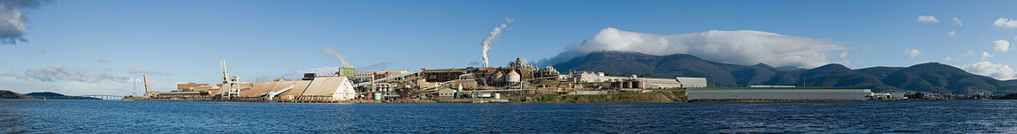 Un panorama que ofrece una gran planta industrial en un lado del mar, en frente de las montañas.