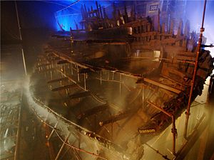Una sección diagonal de un barco de madera visto desde la popa dentro de un edificio moodily iluminado mientras está siendo rociado con agua de un sistema de rociadores