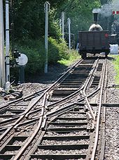 Una vía férrea aleja en la distancia, donde un tren de vapor se encuentra; la pista tiene tres carriles, el medio de que se compensa a la derecha en el primer plano, pero cambia a la izquierda en el medio en algún puntos de presión complejo donde otros tres carriles se unen a la izquierda