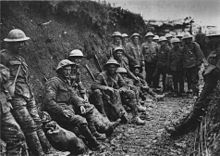 Foto en blanco y negro de dos docenas de hombres con uniformes militares y cascos metálicos sentado o de pie en una zanja fangosa.