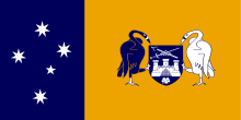 Left third: White stars of the Southern Cross on blue field. Right two-thirds; black and white swans holding a blue shield with a white castle above a white flower and below a sword and scepter arranged in an