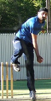 Hombre con barba negro con turbante negro, con pantalones de chándal azul marino, camiseta azul, sigue a través después de una bola de bolos en las redes de cricket. Su pierna izquierda es perpendicular al suelo, y su pierna derecha se eleva y doblada hacia atrás en la rodilla. Su brazo derecho de bolos ha girado a su lado después de soltar la pelota.