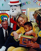 La misma mujer lee un libro en un aula a un niño afroamericano en su regazo, como una niña afroamericana y dos adultos mirad