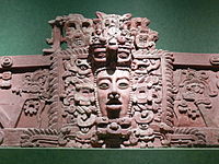 Sección de friso de estuco con rostro humano prominente en el centro, rodeado de elaborada decoración.