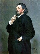 Un homme d'âge moyen avec des cheveux noirs mi-longs et une barbe, vêtu d'un costume sombre, une main dans la poche de son pantalon et l'autre main sur son menton