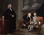 pièce dans les huiles de conversation: homme vêtu de noir avec un col romain se trouve à côté de deux garçons assis sur un canapé, une vêtu d'un costume gris de l'autre un bleu. L'homme tient une feuille de papier; les garçons tiennent un livre.