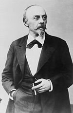 Un homme d'âge moyen avec une calvitie mustachhe et petite barbe, vêtu d'un costume sombre et tenant une cigarette.