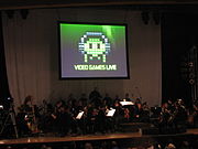 Une photographie d'un orchestre sur une scène faiblement éclairée. Au-dessus du groupe est un écran de projection avec une image en noir, blanc et vert du pixel art. Le pixel art est un objet ovale portant un casque avec des yeux et quatre tentacules. Ci-dessous le pixel art est l'expression