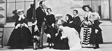 Photo d'une Victoria assis, vêtu de noir, tenant un nourrisson avec ses enfants et Prince Albert debout autour d'elle.