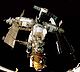 Une station spatiale composée de trois modules blanches disposées en forme de T autour d'un noeud central d'amarrage. Le module sur la course descendante du T a un plus petit module d'orange fixée à l'extrémité distale de celui-ci, et un cinquième module projets vers l'arrière depuis le noeud d'ancrage, avec un sixième module fixé à son extrémité. Un progrès et Soyouz sont à quai à la station, et chacun des modules divers projets solaires plumeuses tableaux, des antennes et des fermes. La noirceur de l'espace constitue la toile de fond de l'image, et les seuils de la soute de la navette spatiale peut être vu en bas de l'image.