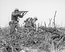 une image en noir et blanc de deux Marines dans leurs uniformes de combat. Marine One fournit couvercle feu avec sa mitraillette Thompson M1 que l'autre avec un fusil automatique Browning, se prépare à briser la couverture de passer à une position différente. Il ya des bâtons et des rochers nus sur le terrain.