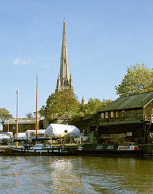 Un clocher d'église sur un quai avec des hangars et des bateaux couverts de bâches en bois. En face de ces derniers sur l'eau un jumeau mat bateau à voile et un narrowboat