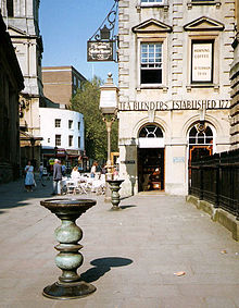 Deux piliers métalliques ornés avec de grands plats sur le dessus dans une rue pavée, avec un bâtiment en pierre du XVIIIe siècle, derrière, sur laquelle on peut voir les mots