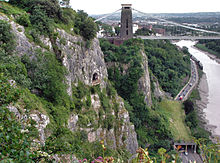 Rocky côté d'une gorge avec une plate-forme en face d'une grotte à mi-chemin. A droite sont une route et la rivière. Dans la distance sont un pont et bâtiments suspension.