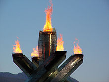 quatre piliers avec la flamme à leurs sommets environnants un seul cinquième pilier au milieu, aussi avec la flamme au sommet. Le fond est ciel avec la montagne.