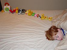 Jeune garçon endormi sur un lit, face à la caméra, avec seulement la tête visible et le corps hors caméra. Sur le lit derrière la tête de l'enfant est une douzaine de jouets soigneusement disposées en ligne.