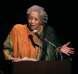 Photo de Toni Morrison, lui montrant dans une robe orange et vert avec ses cheveux gris tirés en arrière. Elle parle à un podium et gestes.