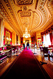 Une photographie d'une grande chambre avec un long tapis rouge qui se étend à travers le milieu de celui-ci et les fenêtres sur le côté droit. Mobilier remplit deux côtés de la chambre. Le plafond contient de moulures et un lustre pend à partir du milieu de l'image.