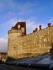 Une photographie d'un haut mur de pierre avec une tour de château sur la gauche, brillant jaune de la faible Sun. La tour et le mur sont percés de petites fenêtres. la tour a un toit de tuiles rouges conique, avec une horloge intégrée dans un côté. Le ciel derrière le bue de mur, avec quelques nuages.