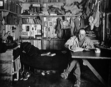 Homme assis les jambes croisées à la table, pipe à la main, apparemment écrit. Beaucoup encombrement des vêtements, des livres et de l'équipement est en arrière-plan.