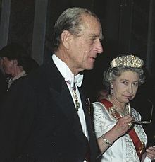 Elizabeth, en robe formelle, détient une paire de lunettes à sa bouche dans une pose réfléchie