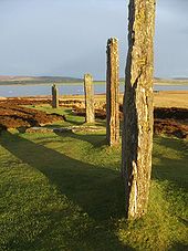 Quatre grandes pierres debout assis dans un champ d'herbe et de bruyère. Ils sont éclairés par la lumière du soleil rougeâtre et ils jettent de longues ombres vers la gauche. Un lac et des collines basses se trouvent au-delà.