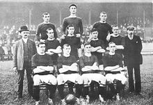 Une photographie en noir et blanc d'une équipe de football aligne avant un match. Quatre joueurs, vêtus de chemises sombres, shorts et chaussettes légères sombres, sont assis. Quatre autres joueurs sont debout juste derrière eux, et trois autres sont debout sur un niveau plus élevé sur la rangée arrière. Deux hommes en costume se tiennent de chaque côté des joueurs.