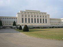 Un lecteur passe devant une pelouse bien entretenue à grand bâtiment rectangulaire blanc avec un colonnes sur elle façade. Deux ailes du bâtiment sont en retrait de la partie centrale.