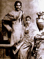 Photographie en noir et blanc d'un homme habillé de manière raffinée et femme: l'homme, souriant, se hanches derrière un régler avec un châle drapé sur ses épaules et en bengali usure formelle. La femme, assise sur le Settle, est en robe et un châle élaboré; elle se penche sur une table sculpté soutenant un vase et les feuilles qui coule.