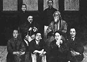 Posé groupe photographie en noir et blanc de sept hommes chinois, peut-être des universitaires, en vêtements de cérémonie: deux portent des costumes de style européen, les cinq autres portent la tenue traditionnelle chinoise; quatre des sept asseoir sur le sol au premier plan; autre est assis sur une chaise derrière eux au centre-gauche; deux autres se tiennent en arrière-plan. Ils entourent un huitième homme qui est vêtu, barbu, et assis dans une chaise placée au centre-gauche. Quatre fenêtres élégantes sont derrière eux en ligne.
