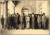 Photo de groupe de plusieurs dizaines de personnes se sont rassemblées à l'entrée d'un bâtiment imposant; deux colonnes dans la vue. Tous les sujets face à la caméra. Tous, sauf deux sont vêtus de costumes de salon: une femme à l'avant-centre porte habit persan de couleur claire; l'homme à sa gauche, première rangée, porte une barbe blanche et chapeau et robe orientale de couleur foncée.