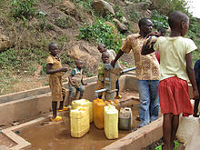Photographie montrant un adulte et cinq enfants de remplissage des jerrycans à une pompe à eau en métal rurale avec socle en béton, au bas d'une colline rocheuse escarpée