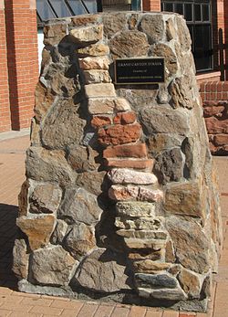 Un pilier solide de la pierre de forme irrégulière motorisée avec des incrustations de empilé plus rock en forme de brique formant une colonne oblique vers la droite. Une plaque sur le pilier lit comme suit: