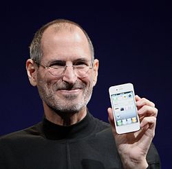 À hauteur d'épaule Portrait de sourire homme dans la cinquantaine porter un col chemise de tortue noire avec une barbe d'un jour tenir un téléphone face au spectateur dans sa main gauche