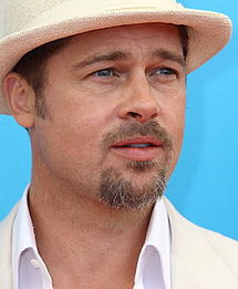 Une race blanche aux cheveux brun clair, les yeux bleus et une barbe brune court, devant un fond turquoise. Il est vêtu d'une chemise blanche et un chapeau blanc.