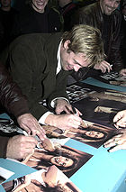 Un homme de race blanche pliée sur une table dédicacer une affiche de film. Il a les cheveux brun clair avec des mèches blondes, et porte un trench-coat de couleur sombre avec une chemise blanche. Visible dans l'arrière-plan et le premier plan sont d'autres personnes, dont certains sont également signer des autographes.