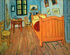 Une chambre étroite avec plancher en bois, murs verts, un grand lit à droite, un deux chaises de paille à la gauche, et une petite table, un miroir et une fenêtre aux volets clos sur le mur du fond. Hanging sur le lit sont plusieurs petites images