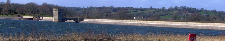 Photo montrant le barrage de couleur blanche se étendant horizontalement à travers l'image avec de l'eau et de la végétation au premier plan et les collines derrière