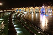 Opinião da noite através de uma praça aberta dominada por uma característica da água longa curva que é decorativamente lit. No outro lado da praça, há a fachada arqueada de um edifício da estação ferroviária