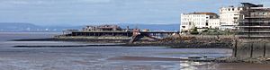 Estrutura com suporte em pernas de metal que saem do mar, que se estende desde a terra que mostra vários edifícios brancos para uma ilha com edifícios nele. Uma casa de barco salva-vidas com rampa de lançamento é anexado ao lado. À distância através da água são colinas e edifícios.
