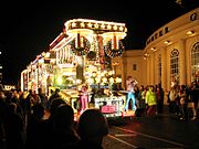 Um grande veículo iluminado por lâmpadas multi-coloridas e transportando pessoas em coustumes brilhantemente coloridos na frente de uma grande multidão em uma rua escura.
