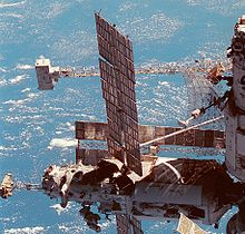 Uma vista parcial de uma estação espacial, com dois módulos visíveis. O módulo tem alinhados verticalmente, uma antena em forma de prato grande que se projecta a partir dele, e um grande fardo pode ser visto horizontalmente por trás dele, com um dispositivo em forma de caixa branca montado na extremidade. O módulo tem uma asna horizontal menor montado na extremidade do mesmo, com uma mochila branco em anexo à presente. O módulo também apresenta dois grandes painéis solares e um número de câmaras instaladas a uma unidade na extremidade distal do mesmo. Outra disposição solar é visível por trás deste módulo.