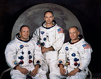 Três astronautas em trajes espaciais sem capacete sentado em frente de uma grande foto da lua.