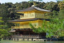 O Pavilhão Dourado é um edifício de três andares com varandas circundando e curvando telhados, com vista para um lago tranquilo e madeiras