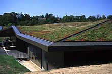 Um baixo edifício tem um telhado completamente coberto com terra e grama. Parece ser construído em uma encosta