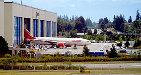 Fábrica de aviões. Edifício rectangular com seis portas, uma das quais é aberto para revelar um avião emergente. O fundo é uma floresta; pequenos veículos e taxiways cercam a fábrica.