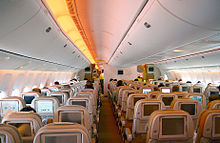 Airliner cabine. Fileiras de assentos dispostos entre dois corredores. Cada encosto tem um monitor; luz brilha a partir das paredes laterais e bagageiros.