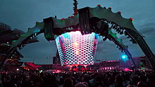 Um palco de concertos; quatro grandes pernas curva-se acima do palco e segure uma tela de vídeo que se estende para baixo para a banda. As pernas são iluminados em verde. A tela de vídeo tem multi-coloridas luzes piscando sobre ele. O público rodeia o palco de todos os lados.
