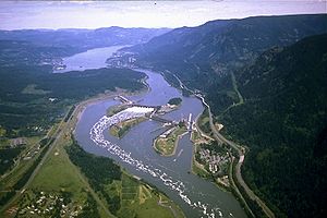 Vista aérea de um grande rio sinuoso através de um desfiladeiro montanhoso. Ele passa sobre uma represa alongamento em quatro segmentos de banco para banco em três ilhas intervenientes. Estradas, passando por conjuntos de edifícios aqui e ali em ambas as margens, correm paralelas ao rio. Whitewater e espuma enrolar rio abaixo de um dos segmentos centrais.