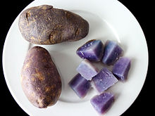 Duas batatas de pele escura em um prato branco. Uma outra batata é cortada em seções para mostrar carne roxo-azul da variedade, colocado no canto inferior direito do mouse sobre a placa.